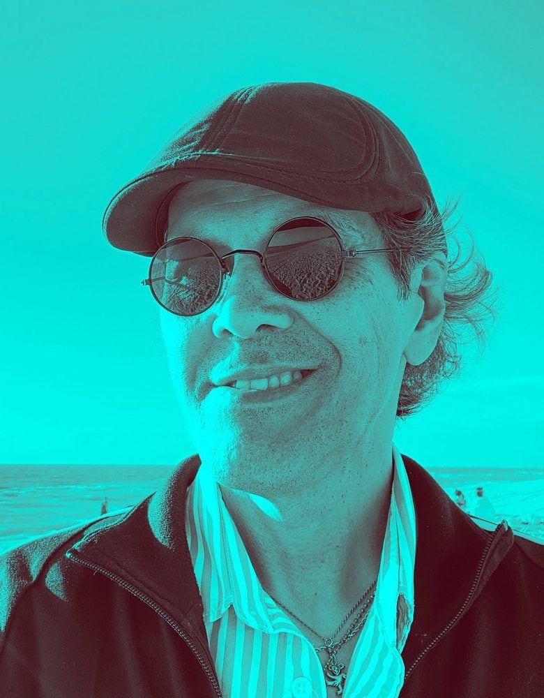 Portrait of Anthony Carroll in a cap and round sunglasses, on a beach