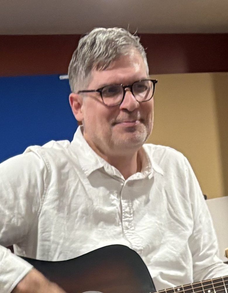 Portrait of Patrick Nolan with an acoustic guitar, seated at a drum kit